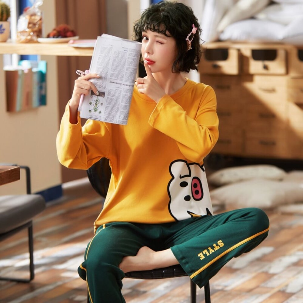 Pigiama di cotone con maglione giallo a forma di maialino e pantaloni verdi, indossato da una donna seduta su una sedia che legge un giornale in una casa