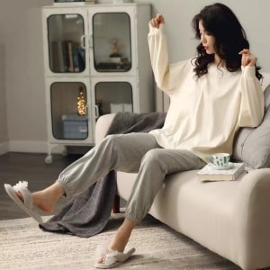 Pigiama estivo con pullover bianco con maniche a pipistrello e pantaloni grigi indossato da una donna seduta sul divano in una casa