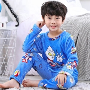 Pigiama di flanella blu da ragazzo supereroe indossato da un bambino seduto su un tappeto davanti a un letto in una casa